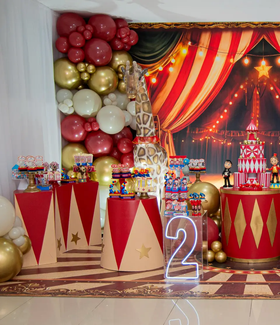 Mesa de festa com tema circo montada no salão da Fest Kids: cortina listrada vermelha, balões dourados e vermelhos e bolo de circo