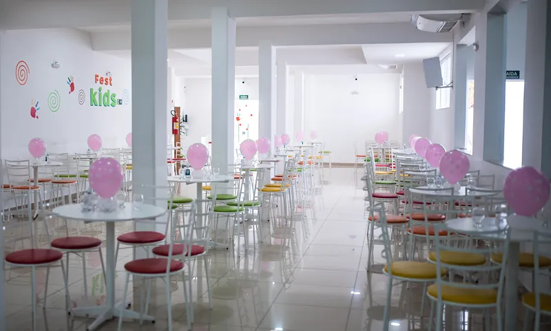 Salão da Fest Kids com mesas redondas brancas, cadeiras coloridas e balões rosa, piso claro e colunas brancas